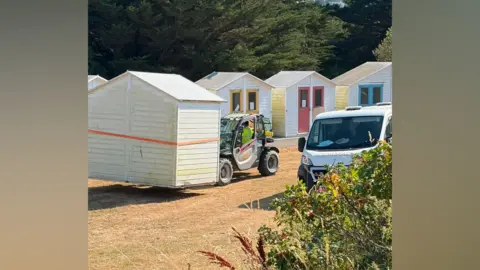 The photo shows a row of timber beach huts on a dry, grassy area, with a small lifting vehicle carrying one of the huts while a white van is parked nearby. Several other huts remain standing in the background, painted white with brightly coloured doors and window frames, while trees form a dense backdrop behind the site.