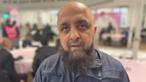 Mahomed Jussab smiles at the camera with a room hall full of people behind him 