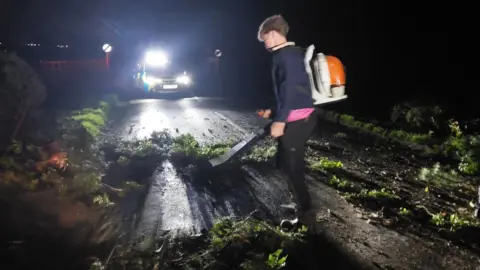 Northamptonshire Police Seamus Durrell with a leaf blower clearing a road. A police car is in the distance, with its lights on. There is a lot of tree debris on the road including leaves and twigs. Seamus is wearing dark trousers, blue top, with a pink top underneath and a leaf blower backpack. 