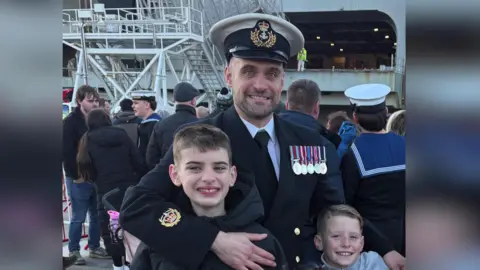 Handout Glen poses for a photograph with his two sons. He is wearing his Royal Navy uniform and the picture appears to have been taken on a naval ship. 