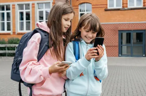 Two school-age girls look at their phones in the playground. They are wearing colourful hoodies and carrying rucksacks on their backs