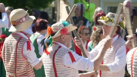 Three Morris dancers in Morris dancing costumes, waving wooden sticks. There is a crowd of people in the background.