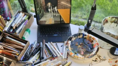 Harriet Lowther Numerous pens and pencils and a paint palette covering a table with a lap top screen in the background displaying an image of a large dog.