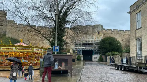 The gate to the castle is held up with scaffolding.  It is raining and a woman walks by with an umbrella.  A man stands looking at the closed gate.