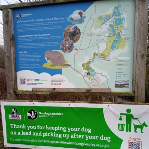 Nottinghamshire Wildlife Trust A wooden information board about the Idle Valley Nature Reserve showing different species and habitats. Below is a green and white banner asking people to clear up after their dogs and to keep their pets on a lead. There is a logo of a person dropping dog mess into a bin and holding a dog on the lead. 