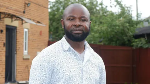 Ngole stands outdoors in a neatly kept garden. He is wearing a light-coloured, long‑sleeved shirt with a subtle floral design and a white undershirt. His hands appear to be resting behind his back. The background includes a well-maintained lawn bordered by small, evenly spaced shrubs. A tall wooden fence runs along the right side of the image, and trees with green foliage are visible behind it. On the left, part of a brick building with white-framed windows and a black door can be seen.