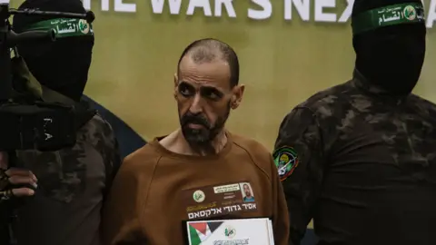 HAITHAM IMAD/EPA-EFE/REX/Shutterstock Eli Sharabi flanked by two Palestinian fighters. He is wearing a brown sweatshirt with a banner containing writing and a small picture of a man on. The men either side of him are wearing black face coverings with a green banner on and are wearing long sleeved tops with dark camouflage and a badge on the arm. A gun can be seen in the left hand side of the photo.