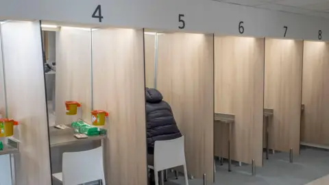 PA Media Several cubicles, numbered from 4 to 8, lined up - there are mirrors at each one, with a chair and other items on each table. A man wearing a dark jacket, with his head not visible is sitting at No 5.