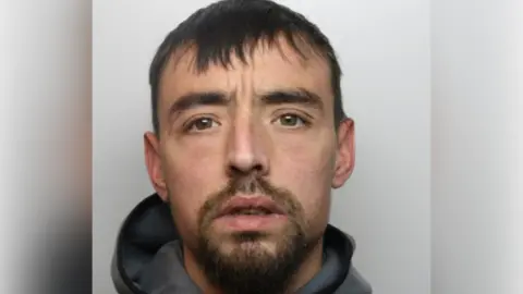 A mugshot of a man with short black hair, a beard and grey hooded top.
