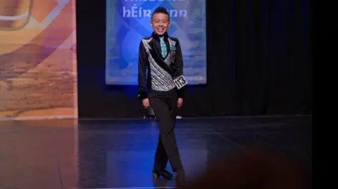 HopSkip Productions A young boy wearing a waistcoat and black trousers, legs crossed dancing, he is smiling on stage