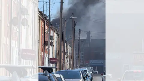 A large plume of black smoke billows across a terraced street.
