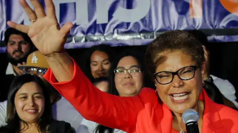Incumbent mayor Karen Bass holds her hand up in a wave as she speaks at a microphone in front of supporters