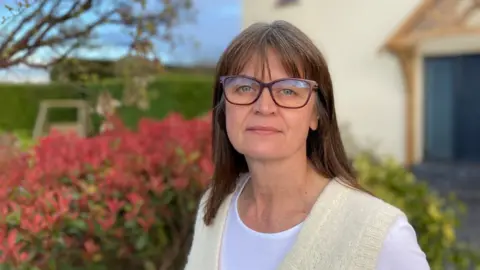 A woman wearing glasses and a white top with a cream-coloured cardigan is standing outdoors. She has brown hair and is looking directly at the camera. Behind her is a garden with red plants and a light-coloured building.