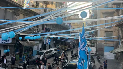 Blue and white ribbons and streamers are stretched high across the streets between buildings on either side of a street. There are also blue balloons and Napoli flags. There are people and cars below them.