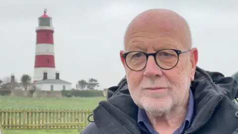 A head and shoulders image of Harry Blathwayt. He is wearing a dark, hooded top and glasses. Happisburgh Lighthouse is fully in view over his right shoulder. It has three red and two white stripes around it, below the lantern, and a cottage at its base.