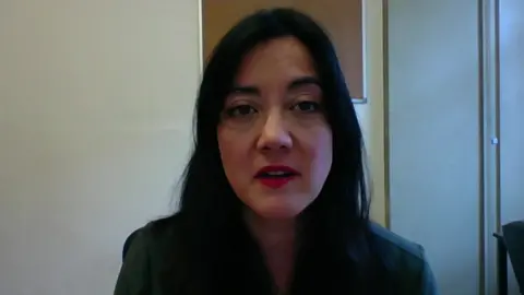 MP Sarah Owen, speaking via computer video communication software. She has long dark hair and wears red lipstick, with an office wall visible in the background.