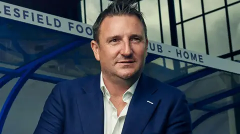 A headshot of Smethurst. He had short brown hair which is spiked at the front, and is wearing a blue jacket and white shirt. He is standing in front of the dugout of the club's stadium.