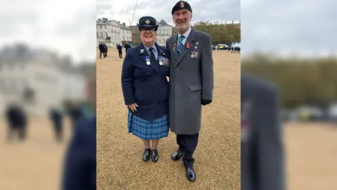 Tracy, who is wearing military uniform and a hat, stood next to her dad, Mel, who is wearing a long grey coat and military badges. It is a full body shot. They are both looking at the camera smiling. 