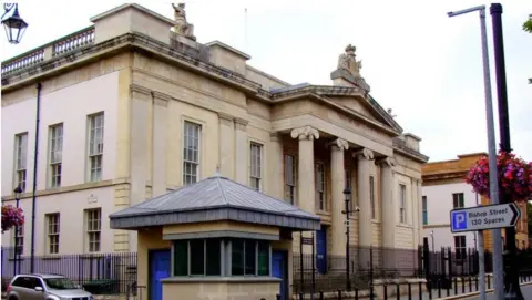 Shows a courthouse with four pillars at the entrance and a security sangar to the left. A silver-coloured car is on the left of the picture and, on the right, a signpost pointing to Bishop Street car park and saying it has 130 spaces.
