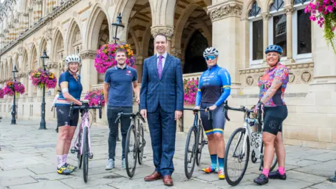 West Northamptonshire Council Man in a blue chequered suit stands between four cyclists in front of the Guildhall 
