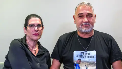 Talik, who has black hair in a ponytail and wears black-rimmed glasses, poses for a photo with her husband Itzik, who has grey short hair and a beard and wears a silver chain. They both have neutral expressions.