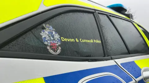 The side of a Devon and Cornwall Police car. The police logo is on display on the window. Rain droplets are falling down the exterior of the car. It has blue and yellow stickers. 