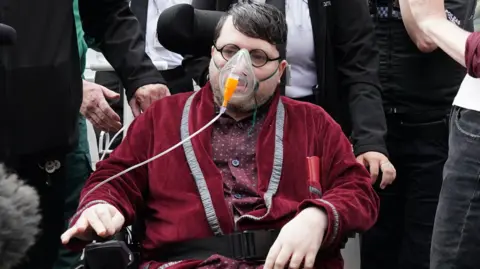 PA Media Nicholas Rossi outside court in purple pyjamas and dressing gown. He is sitting in an electric wheelchair and is wearing an oxygen mask and glasses.