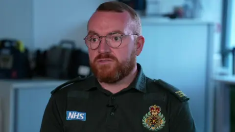 BBC James Vernon, who has short ginger hair, a ginger beard and round framed glasses, sits on a chair and speaks to the camera wearing his green North West Ambulance uniform. 
