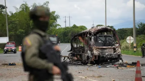 A solider in the foreground holds a gun against a backdrop of a burnt-out vehicle