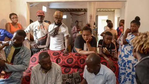 AFP via Getty Images A family group look mournful as they stand behind a coffin wrapped in a red patterned cloth.