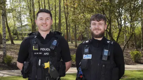 Police Federation PC Sam Clover and PC Jacob Edwards standing outside next to each other. They are both wearing black police uniform and are looking directly at the camera and smiling. There are trees behind.