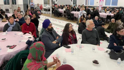 people from different faiths sit at tables in a large hall looking towards a stage 