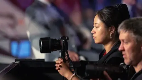 Yu, a Chinese woman in her mid twenties in this picture, looking through the lens of a camera to the left hand side of the frame. She has a neutral, focused expression. She has quite tanned skin, chunky earrings, and black hair tied up in a clip.