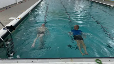 A boy is swimming backstroke next to his swimming teacher. he has his head tilted back and is wearing goggles. His teacher, a man, is wearing a blue t-shirt and black shorts.
