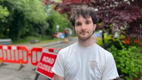 Ross MacPhee standing on a residential street in front of red “Road Closed” safety barriers, with trees and greenery in the background. He is wearing a light-coloured T-shirt and is positioned in the foreground, with roadworks equipment and signage visible behind.