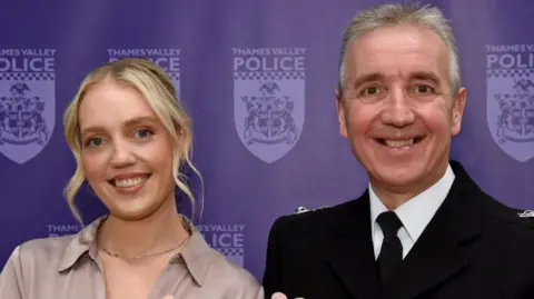Victoria and Jim Weems. Victoria has medium-length blonde hair and is wearing a bronze blouse, whist Jim stands next to her in official police dress. Behind the pair is a blue wall with the Thames Valley Police logo printed on it.
