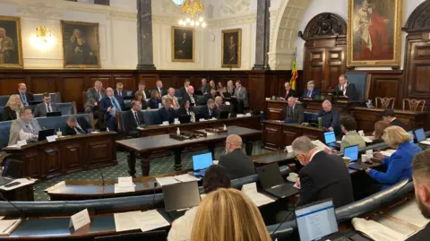 A council chamber full of councillors sat down in a meeting.
