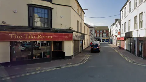A street view of Rhyl town centre, with The Tiki Room on the left. The yellow building is on a street corner with a red sign running along it with the name 'The Tiki Room'. A doorway and two large windows are seen below with a bay window on the first floor. 