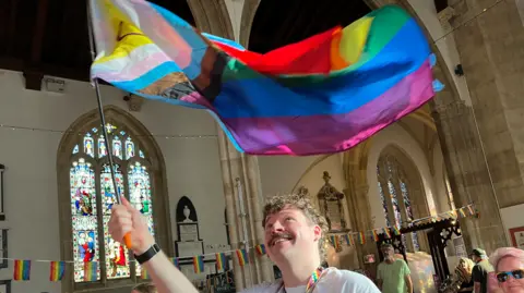 A man waving a Pride flag inside a church. The man, who has blonde, curly hair and a moustache, is looking up at the flag and smiling. There are people gathered behind him.