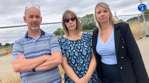 LDRS A bald man with a grey beard wearing a blue and grey striped t-shirt. He has his arms crossed and stands next to two women - one with brown hair and wearing sunglasses and a blue dress, the other with blonde hair and wearing a black jacket and blue top. They are all looking at the camera, there is a metal fence behind them.