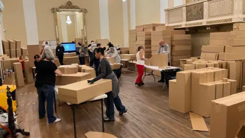 People are using cardboard to create various structures which will be part of the finished work. Many have gloves and they are working in pairs. The large room is full of stacked-up boxes.