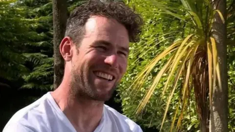 BBC Mark Cavendish smiling. he's wearing a white T-shirt and sitting in a garden with green trees behind him.