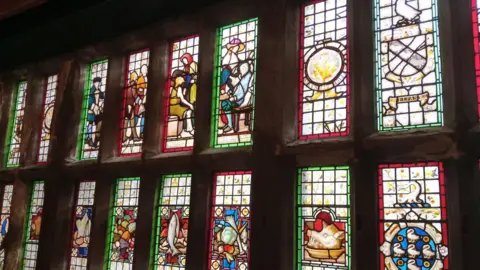 Tony Callahan A row of stained-glass windows inside a historic building, illuminated by light shining through multi-coloured panels. The windows feature decorative illustrations, including figures, animals, heraldic-style symbols and ornamental designs framed in red, green and yellow glass. Dark wooden surrounds separate the panels, and the warm interior lighting highlights the rich colours and intricate details of the artwork.