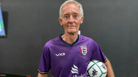 BBC Geoff Martel is wearing a purple shirt with the England logo which features three blue lions. He is holding a white football with green lines on it under his left arm. he has short white hair and is looking straight at the camera.