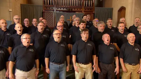 Man stood in navy polo shirts singing inside a church with an organ in the background. They are all different ages.