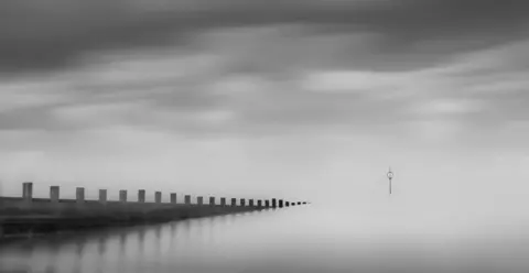 A black‑and‑white scene showing a long wooden pier stretching diagonally into calm, misty water. The sky is cloudy and blurred, and a single navigation marker stands alone in the distance.