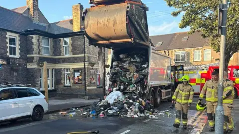 Refuse strewn on the road in the aftermath of a refuse wagon blaze with firefighters and a fire engine also in view