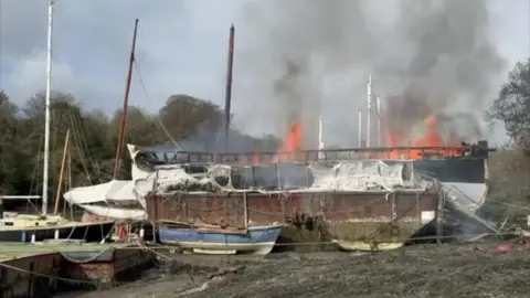 A picture of the fire which has taken over the boat in the estuary. There is large orange flames.