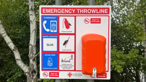 A wide-shot image of an emergency throwline station. It is bright blue, orange and red. There are green trees behind.