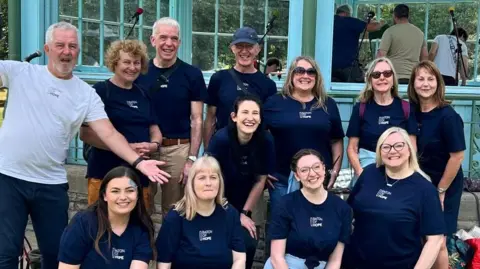 Ten people in navy blue T shirt and one man in a white T shirt pose in front of a teal bandstand. They are smiling at the camera.